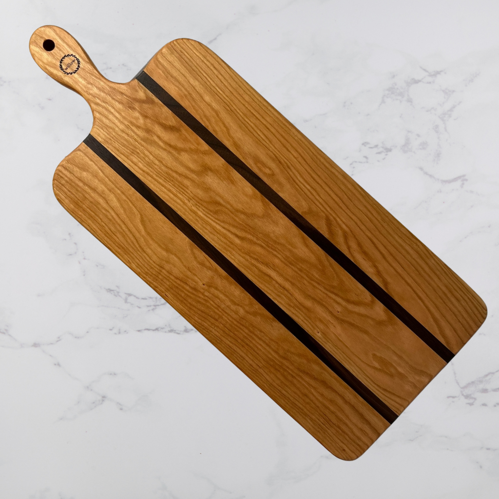 A long charcuterie board made of Vermont cherry with black walnut accents, placed on a marble surface.
