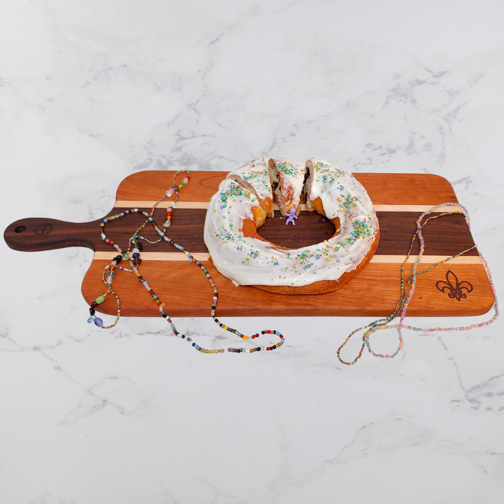 King cake on a wooden charcuterie board engraved with a Fleur-de-lis with colorful Mardi Gras beads on a marble surface